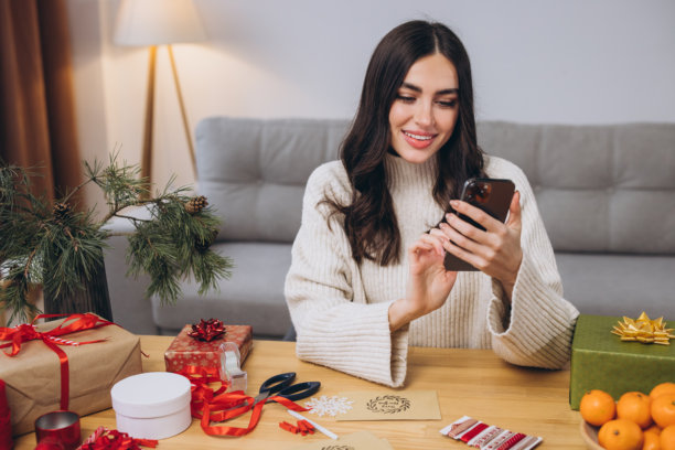 快乐的女人在家中使用智能手机，身边是礼物和模糊的装饰。图片下载