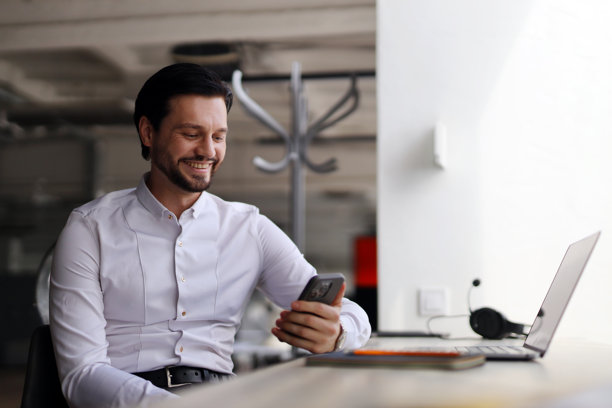 一个男人坐在桌子前，面前有一台笔记本电脑和一部手机。他面带微笑，正在看着他的手机。图片下载