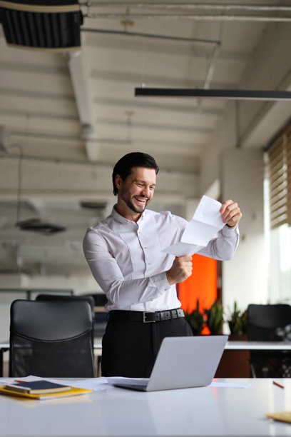 男人面带微笑，手拿一张纸，房间里有笔记本电脑和椅子图片下载