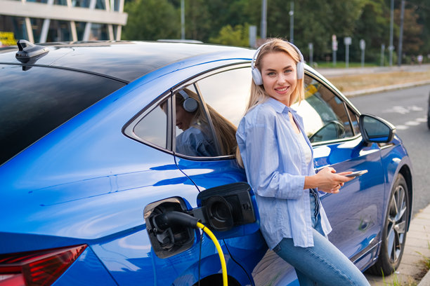 年轻女人戴着耳机放松，在她的电动汽车充电时查看手机图片下载