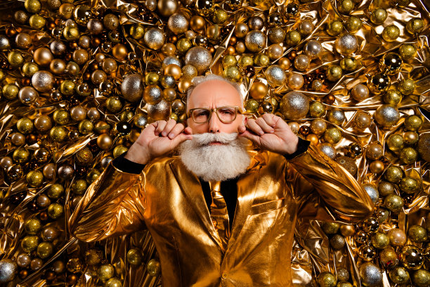 俯视照片中，一位酷炫的老年男子躺在圣诞树饰品上，轻抚着修剪整齐的胡须，穿着光亮时尚的服装。图片下载