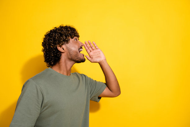侧面照片，快乐的男孩，波浪形头发，穿着卡其色T恤，朝着空旷的销售空间大喊，背景为黄色。图片下载