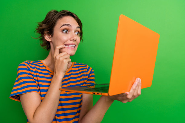 漂亮年轻女孩的照片肖像，手持笔记本电脑，咬着指甲，表现出兴趣，穿着时尚的条纹服装，背景为绿色。图片下载