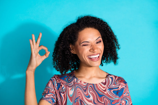 漂亮年轻女孩的照片肖像，做出“OK”手势，眨眼，穿着时尚多彩的衣服，背景为水绿色。图片下载