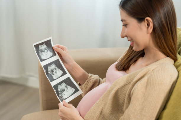 快乐的年轻孕妇在家中看着超声波扫描照片，抚摸着自己的肚子，期待着幸福的母亲时光，准备迎接孩子的出生，进行家庭规划并为孩子的健康投保。图片下载
