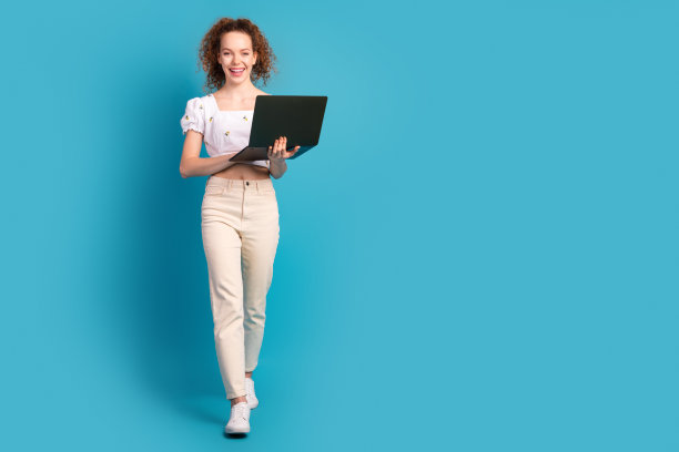 穿着时尚白色服装的漂亮年轻女孩在蓝色背景下走路，手里拿着笔记本电脑的全身照片图片下载