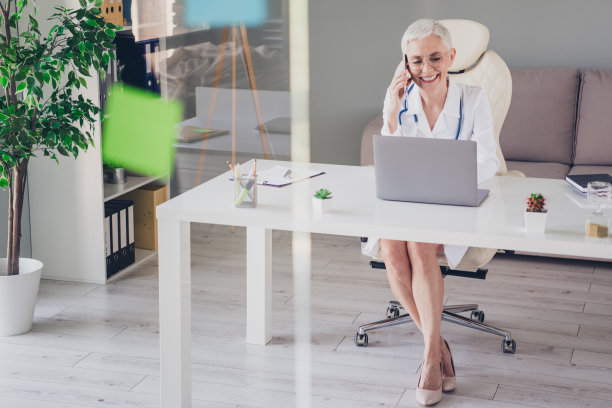 全身照片，年长女性合格医生在办公室室内穿着白大褂，使用手机和笔记本电脑。图片下载