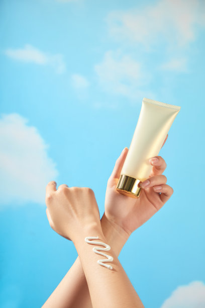 模特的手相互摩擦，挤出一条锯齿状的奶油，手握着一个带有铜金色盖子的米色管状奶油样品，背景是蓝色的夏季场景，用于防晒广告照片模板。图片下载