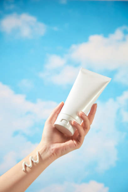 在这张用于宣传护肤产品的照片中，背景是美丽的蓝天和飘动的云朵。一位女性模特正手持一支无品牌的白色管子，手腕上涂抹着一抹奶油。图片下载