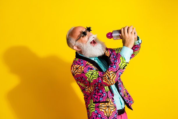 时尚的老年男性穿着华丽的粉色服装，戴着太阳镜，独自一人在黄色背景下唱卡拉OK的肖像照片图片下载