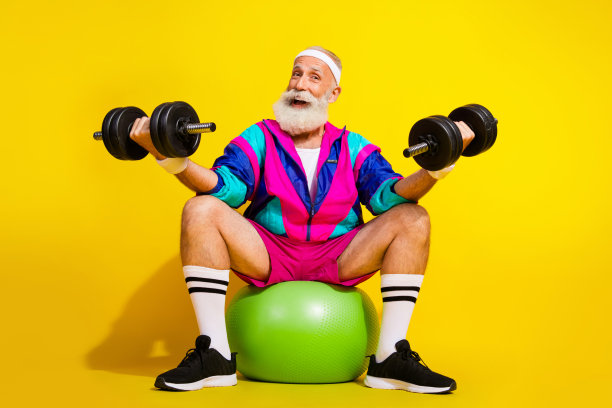 兴奋的运动员爷爷穿着时尚复古运动服，举着哑铃和健身球，背景为黄色，完整尺寸的照片海报。图片下载