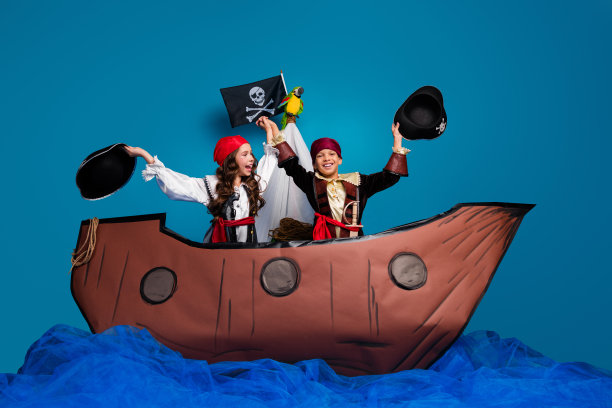 两位小朋友穿着海盗服装，举起帽子庆祝生日派对，正在蓝色背景下玩船的全身照片。图片下载