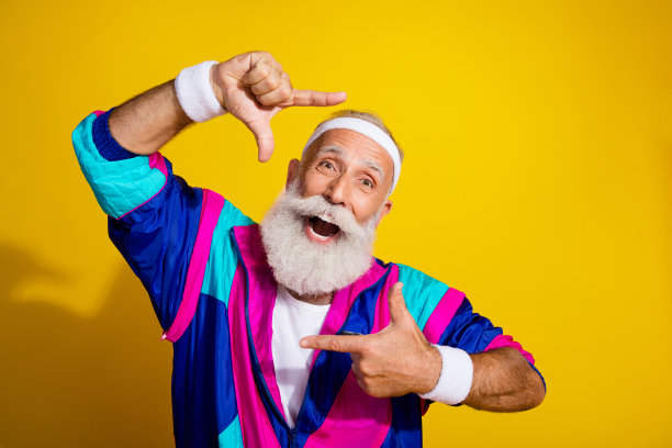 穿着复古运动夹克的快乐成熟男士，做出展示照片的手势，背景为空旷的黄色。图片下载