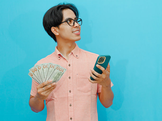 微笑的亚洲商人手握现金和手机，抬头思考，策划某事。背景为蓝色，适合商业或金融概念的图片。图片下载