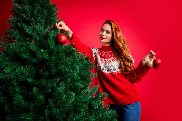 穿着节日毛衣的年轻女孩在红色背景下挂着装饰球和玩具，身旁是常青树的照片肖像。图片下载