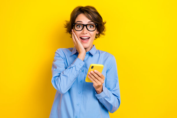 特写照片：年轻迷人、有趣、兴奋、积极、惊讶的女性，捂着脸颊，手持手机，正在进行夏季促销的网上购物，背景为黄色。图片下载