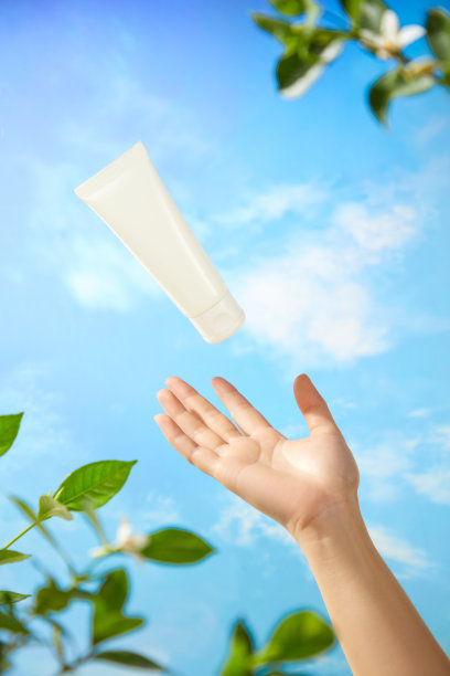 一张用于宣传美容产品的照片，展示了一位模特的手伸向一支未标记的白色乳霜管，背景是美丽的蓝天，漂浮着白云和一些树枝。图片下载