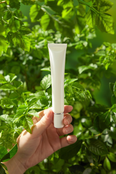 这张图片非常适合用于天然美容护理产品的营销，因为它展示了一只模特的手轻柔地握着一支白色护肤霜管的模型，背景是模糊的植物景观。图片下载