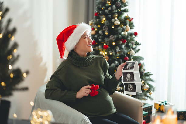 亚洲孕妇戴着圣诞老人帽，开心地微笑，手里拿着超声波照片和钩织的婴儿鞋，坐在装饰有圣诞树的客厅里。图片下载