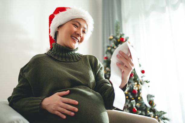 亚洲孕妇戴着圣诞老人帽，开心地微笑，手中拿着超声波照片，坐在装饰有圣诞树的客厅里。图片下载