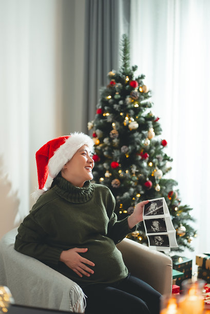 亚洲孕妇戴着圣诞帽，开心地微笑，手中拿着超声波照片，坐在装饰有圣诞树的客厅里。图片下载