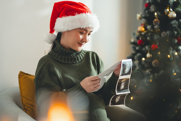 亚洲孕妇戴着圣诞老人帽，开心地微笑，手中拿着超声波照片，坐在装饰有圣诞树的客厅里。图片下载