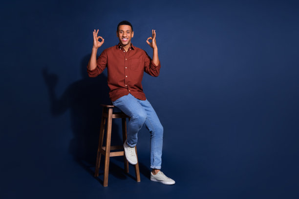 穿着棕色衬衫的开朗积极的家伙坐在凳子上的全长照片，显示出空白的空间孤立的蓝色背景图片下载