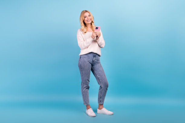 照片中，开朗可爱的女人穿着时髦的白色衣服，拿着蓝色背景的手机图片下载