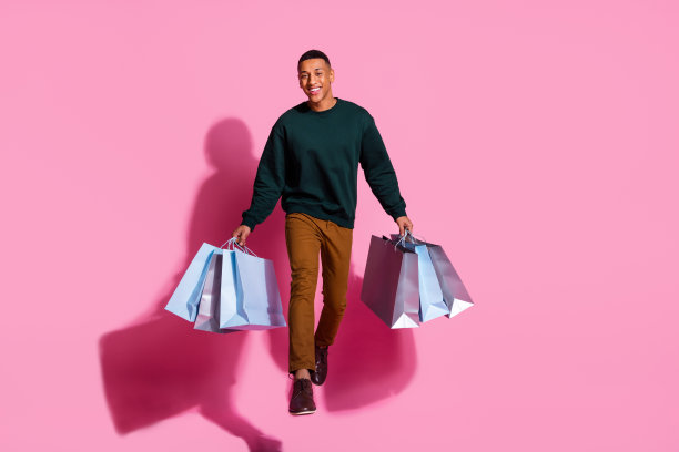 全身照片英俊的好心情的家伙穿着运动衫跳抱购物者空旷的空间孤立的粉红色背景图片下载
