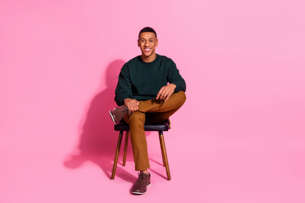时髦酷的家伙穿着套头衫坐在扶手椅上微笑的全身照片，空旷的空间孤立的粉红色背景图片下载