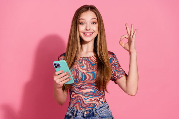 照片中，满意的女孩直发，穿着印花t恤，拿着智能手机，在粉红色的背景上显示出ok的认可图片下载