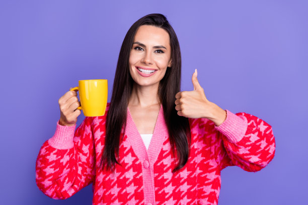 照片中可爱可爱的女孩穿着粉红色的开襟羊毛衫，大拇指向上翘着，享受咖啡，空旷的空间孤立的紫色背景图片下载