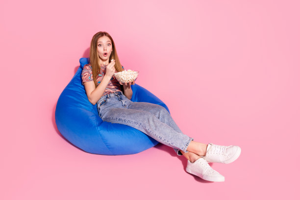 可爱的少女女士坐豆袋吃爆米花电影震惊穿着时尚的印花衣服孤立在粉红色的背景照片图片下载