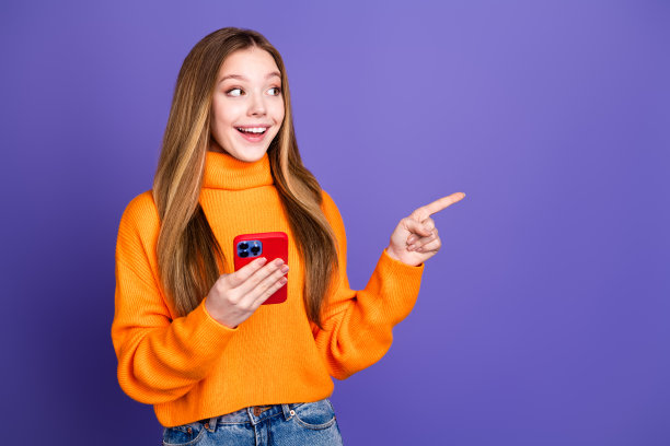 照片的青年金发可爱的快乐的微笑女孩在橙色高领毛衣使用智能手机推荐酷的应用程序孤立的紫罗兰色背景图片下载