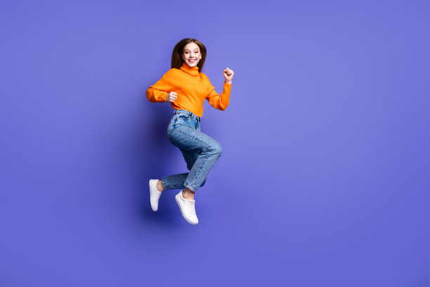 照片肖像金发开朗的年轻女孩在橙色高领毛衣和牛仔裤跑快练习运动孤立的紫色背景图片下载