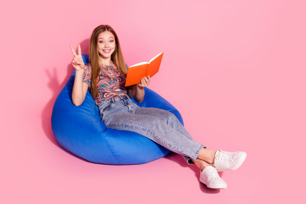 可爱的十几岁的女士坐豆袋阅读书籍显示v字穿着时尚的印花衣服孤立在粉红色的背景照片图片下载