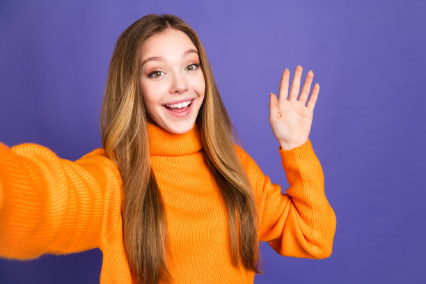 自拍照片的青年迷人的金发可爱的快乐的微笑女孩在橙色毛衣挥手进入干部孤立的紫色背景图片下载