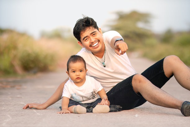 父亲和婴儿男孩坐着，指着镜头。他们穿着白侣装。照片的氛围是在阳光明媚的下午户外。图片下载