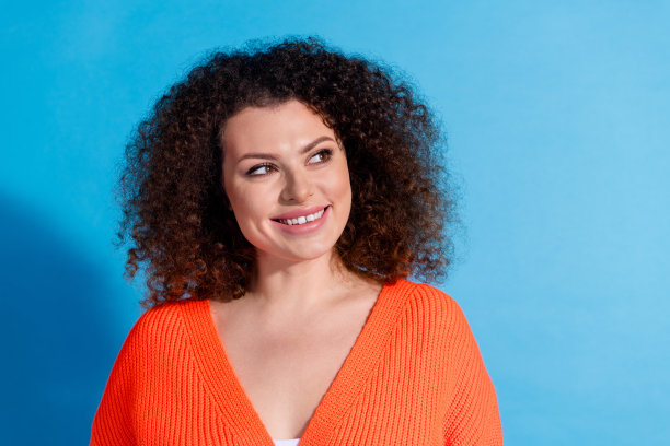 穿着橙色开衫的波浪卷发模特女性在蓝色背景前注视新系列的照片肖像图片下载