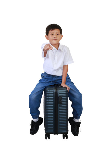 亚洲小男孩骑在行李箱上，指着镜头，直视前方，背景为白色。儿童与旅行的概念。全身照片，带有抠图路径。图片下载