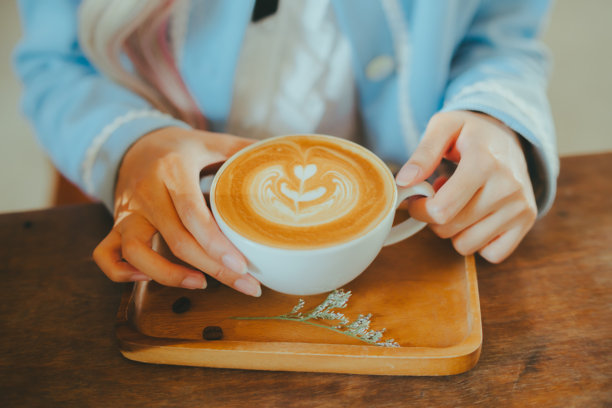 一张特写照片，展示一位女性的手握着一杯咖啡，上面有拉花，放在木桌上。图片下载