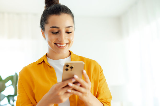 使用电子设备。照片中是一位穿着橙色衬衫的积极美丽的阿拉伯或印度年轻女性，手中拿着智能手机，正在社交媒体上发送消息、浏览互联网和查看电子邮件。图片下载