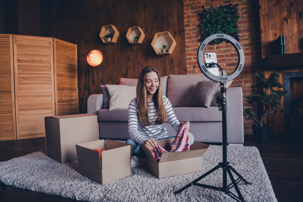 可爱年轻女孩在现代阁楼公寓室内拆包衣物，三脚架支撑智能手机录制视频的全景照片图片下载
