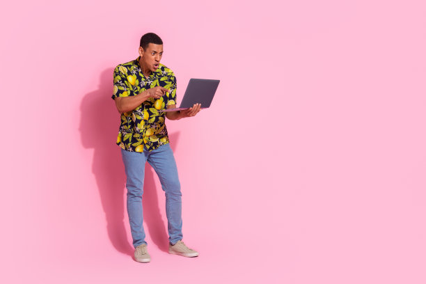 年轻男性在粉色背景下穿着时尚的热带印花服装，愤怒地通过视频通话抱怨，手持笔记本电脑的全身照片。图片下载