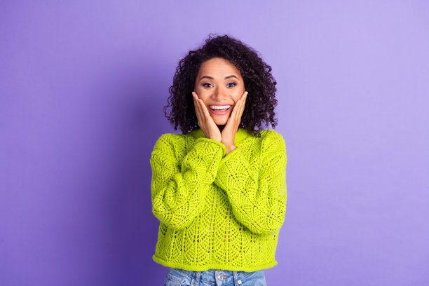 紫色背景下，穿着绿色时尚针织毛衣的可爱女学生，波浪卷发，惊讶地捂着脸颊的肖像照片。图片下载