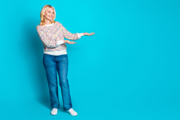 穿着毛衣的优雅老年女性在青色背景前手持空白广告牌的全身照片图片下载