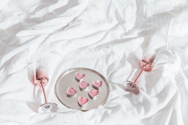 两只粉色闪亮的香槟杯和燃烧的心形蜡烛摆放在白色床上。极简生活美学照片，情人节，浪漫关系，爱情概念，浪漫约会。图片下载