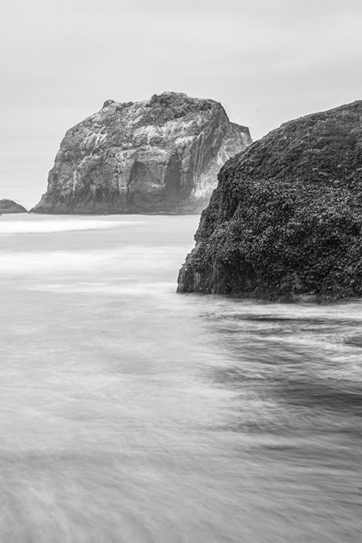一张长时间曝光的黑白照片，拍摄于俄勒冈州班登的海滩，天空多云。长时间曝光模糊了海浪的运动。图片下载