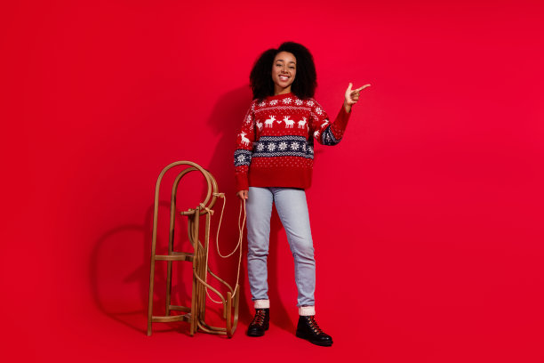年轻女士穿着圣诞服装，手持雪橇指向空白处，在红色背景下庆祝冬季假期的全身照片。图片下载