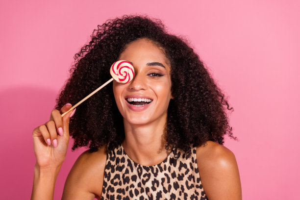 可爱的年轻女士的照片肖像，手持棒棒糖遮住眼睛，穿着时尚的豹纹服装，背景为粉色。图片下载